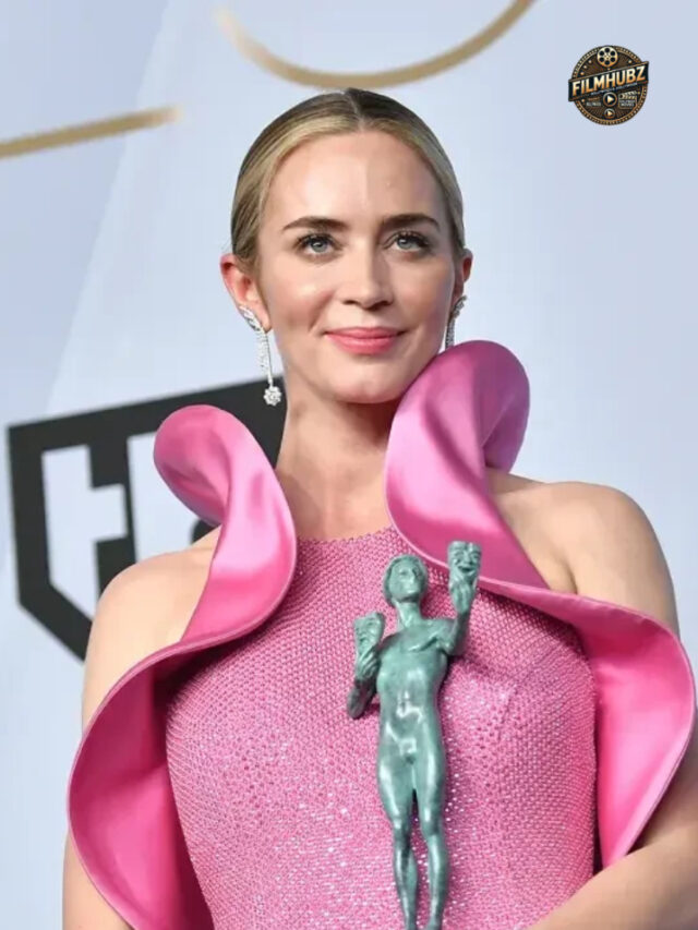 Emily Blunt smiling in an elegant dress, speaking at a gala event.
